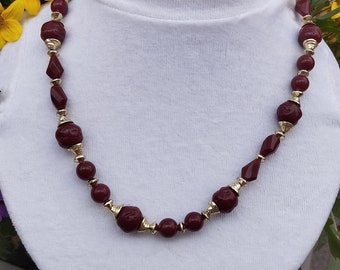 May include: A maroon and gold beaded necklace with a variety of shapes and sizes of beads. The necklace is displayed on a white t-shirt.