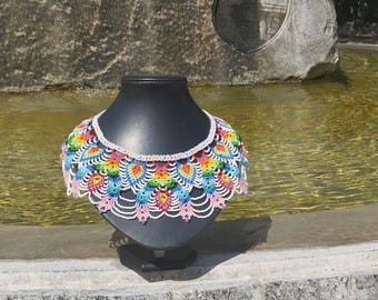 May include: A colourful beaded necklace with a rainbow pattern. The necklace is made of small, white beads with colourful accents. It is displayed on a black mannequin against a background of stone and water.