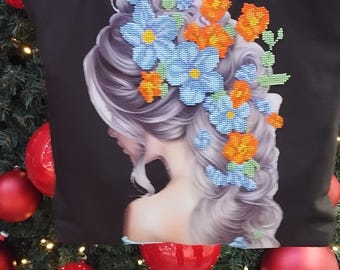 May include: A black tote bag featuring a design of a woman's profile with blue and orange flowers in her hair. The bag is positioned in front of a Christmas tree adorned with red baubles.