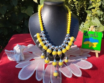 May include: A yellow beaded necklace with silver details and hanging pendants. The necklace is displayed on a black mannequin head. A small painting of the Ukrainian flag is visible. The background is red.