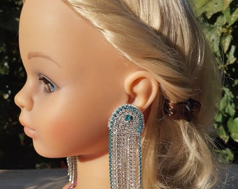 May include: A pair of long, silver dangle earrings with blue crystal accents. The earrings are made of a chain of small, clear crystals with a larger, blue crystal at the top.