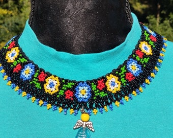 May include: A black beaded choker necklace with a colourful floral pattern and a blue glass pendant with silver angel wings. The necklace is on a turquoise shirt.