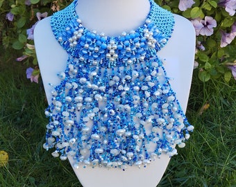 May include: A blue and white beaded necklace with a fringe of beads. The necklace is on a white mannequin bust and is set against a background of green grass and purple flowers.