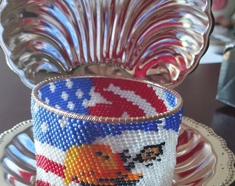 May include: A beaded bracelet featuring an American flag design and an eagle in the centre. The bracelet is displayed on a silver tray with a shell-shaped lid.