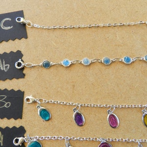 Puede incluir: Cuatro pulseras de cadena plateadas con diferentes dijes. La pulsera superior tiene un dije azul con una imagen religiosa. La segunda pulsera tiene varios dijes azules. La tercera pulsera tiene cuatro dijes de vidrio de diferentes colores. La pulsera inferior tiene varios dijes de vidrio de diferentes colores.