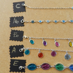 Puede incluir: Cinco pulseras de cadena plateadas con diferentes dijes. Los dijes son de forma ovalada y presentan una imagen azul, verde, roja, morada y amarilla de la Virgen Mar&iacute;a. Las pulseras est&aacute;n etiquetadas con A, B, C, D y E.