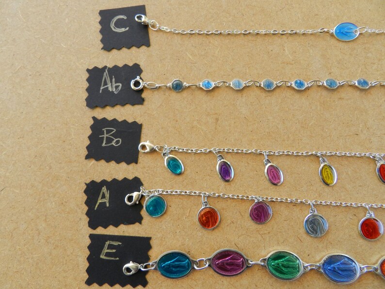 Puede incluir: Cinco pulseras de cadena plateadas con diferentes dijes. Los dijes son de forma ovalada y presentan una imagen azul, verde, roja, morada y amarilla de la Virgen Mar&iacute;a. Las pulseras est&aacute;n etiquetadas con A, B, C, D y E.