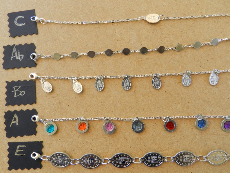 Puede incluir: Cinco pulseras de cadena plateadas con diferentes dijes. Los dijes incluyen peque&ntilde;os c&iacute;rculos plateados, medallones ovalados con un dise&ntilde;o de cruz y piedras de vidrio de colores.