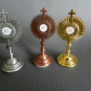 Puede incluir: Tres pequeños soportes religiosos decorativos con un diseño de cruz y sol. Los soportes están hechos de metal y están acabados en plata, cobre y oro. Cada soporte presenta una placa circular blanca con las letras "JHS" en el centro.
