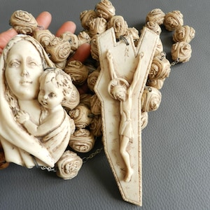 Può includere: Rosario color avorio con perline scolpite e ciondoli religiosi. I ciondoli raffigurano la Vergine Maria con il bambino Gesù e un crocifisso con Gesù Cristo. Le perline sono di colore beige chiaro e lo sfondo è grigio neutro.