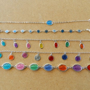 Puede incluir: Un conjunto de nueve pulseras de plata con abalorios de colores. Cada pulsera presenta un abalorio de color diferente con una imagen religiosa. Los abalorios son azules, azul claro, morados, rosas, naranjas, rojos, verdes, amarillos y turquesas.