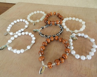 Puede incluir: Seis pulseras diferentes con dijes religiosos. Las pulseras están hechas de cuentas blancas, cuentas de madera marrón y cuentas negras y blancas.