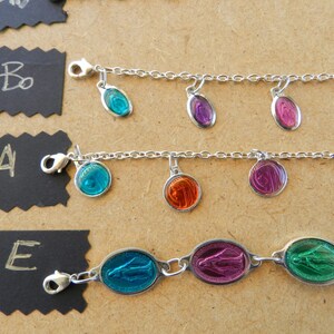 Puede incluir: Tres pulseras de plata con diferentes dijes de vidrio de colores. Los dijes tienen forma ovalada y presentan una imagen religiosa. Las pulseras est&aacute;n sobre una superficie marr&oacute;n con etiquetas negras.