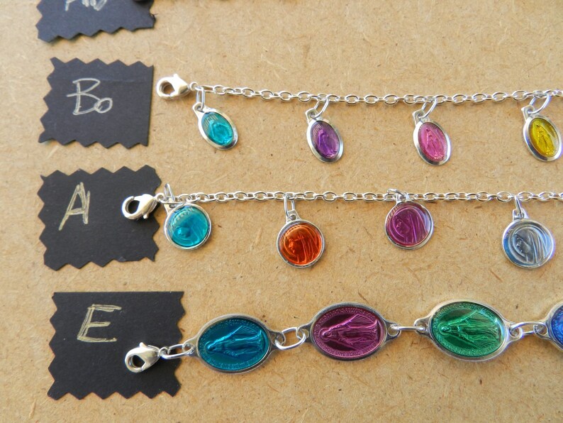 Puede incluir: Tres pulseras de plata con diferentes dijes de vidrio de colores. Los dijes tienen forma ovalada y presentan una imagen religiosa. Las pulseras est&aacute;n sobre una superficie marr&oacute;n con etiquetas negras.