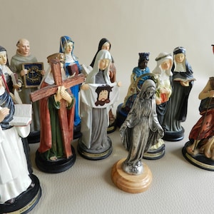 Op de afbeelding: Een verzameling van 12 religieuze beeldjes, elk met een andere heilige. De beeldjes zijn gemaakt van geverfd hars en staan op individuele voetstukken. De heiligen zijn onder andere Sint Franciscus, Sint Antonius, Sint Jozef, Sint Maria, Sint Petrus, Sint Paulus, Sint Johannes de Doper, Sint Michaël de Aartsengel, Sint Gabriël de Aartsengel, Sint Rafaël de Aartsengel, Sint Judas Thaddeus en Sint Christoffel.