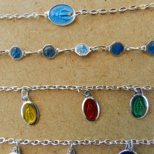 Puede incluir: Cuatro pulseras de cadena de plata con diferentes dijes. La pulsera superior tiene un dije ovalado azul con una figura religiosa. La segunda pulsera tiene cinco dijes octogonales azules con figuras religiosas. La tercera pulsera tiene cuatro dijes ovalados con figuras religiosas de diferentes colores. La pulsera inferior tiene cuatro dijes ovalados con figuras religiosas de diferentes colores.