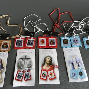 Peut inclure: Cinq scapulaires différents avec des images religieuses. Chaque scapulaire a une paire de petites étiquettes rectangulaires en tissu avec des images de Jésus, Marie et le Sacré-Cœur. Les scapulaires sont numérotés de 1 à 5. Les scapulaires sont sur une surface grise.
