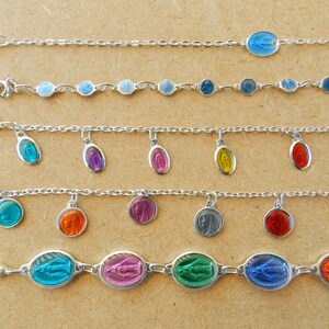 Puede incluir: Cinco pulseras de cadena plateadas con diferentes encantos ovalados de colores. Los encantos presentan una imagen religiosa. Las pulseras est&aacute;n sobre una superficie marr&oacute;n.