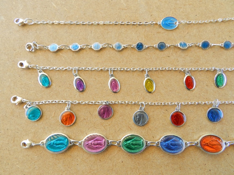Puede incluir: Cinco pulseras de cadena plateadas con diferentes encantos ovalados de colores. Los encantos presentan una imagen religiosa. Las pulseras est&aacute;n sobre una superficie marr&oacute;n.