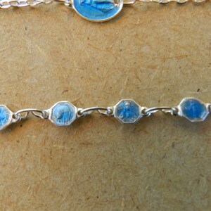 Puede incluir: Un brazalete de cadena de plata con dijes de esmalte azul que representan figuras religiosas. Los dijes son de forma octogonal y tienen un borde plateado.