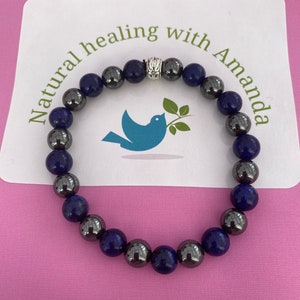 Puede incluir: Un brazalete hecho de cuentas de color azul oscuro y negro con un dije de plata. El brazalete está sobre una superficie rosa con una tarjeta blanca que dice "Natural healing with Amanda".