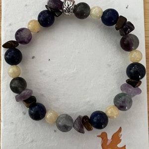 Può includere: Un braccialetto di perline con una varietà di pietre, tra cui ametista, lapislazzuli, occhio di tigre e fluorite. Il braccialetto ha un ciondolo d'argento con un motivo floreale.