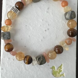 Op de afbeelding: Een armband gemaakt van verschillende stenen, waaronder tijgeroog, helder kwarts en grijze stenen. De armband heeft een zilveren sluiting met een Grieks sleutelontwerp.