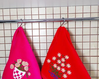 May include: Two kitchen towels hanging on a rack. The towel on the left is pink with a floral design and a checkered pot. The towel on the right is red with a white daisy design.