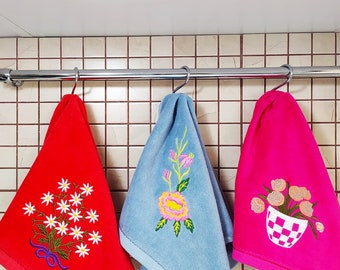 May include: Three embroidered kitchen towels hanging on a silver rack. The towels are red, blue, and pink with floral designs.