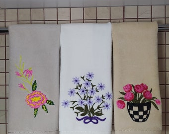 May include: Three embroidered kitchen towels hanging on a rack. The towel on the left has a pink and green floral design. The middle towel has a purple and green floral design. The towel on the right has a pink flower design in a black and white checkered pot.