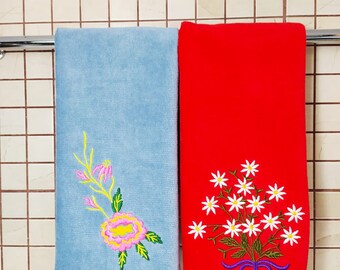 May include: Two embroidered hand towels hanging on a rack. One towel is light blue with a pink and yellow flower design. The other towel is red with a white flower bouquet design.