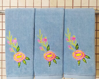 May include: Three light blue towels with embroidered pink and yellow flowers. The towels are hanging on a rack.