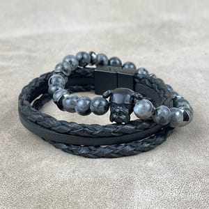 Puede incluir: Un brazalete de cuero negro con un diseño trenzado y un brazalete de cuentas de piedras preciosas negras con un dije de bulldog negro.