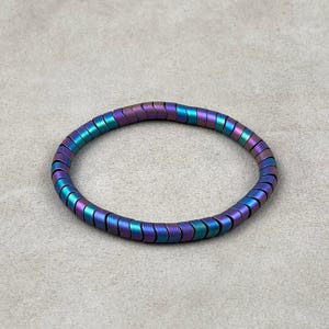 May include: A multi-colored bracelet with a metallic sheen. The bracelet is made of small, rectangular beads that transition from purple to teal and blue. The bracelet is circular and sits on a neutral gray surface.
