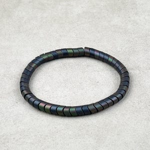 May include: A dark, iridescent bracelet composed of small, curved, rectangular beads. The beads have a metallic sheen, displaying hues of blue, green, and purple. The bracelet is circular and rests on a neutral, textured surface.