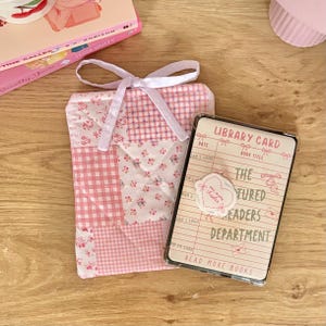 May include: A quilted fabric book sleeve featuring a pink and white patchwork pattern of gingham and floral prints, tied with a white ribbon. Beside it lies a rectangular clear plastic card holder displaying a library card design with the text "LIBRARY CARD," "THE FEATURED READERS DEPARTMENT," and "READ MORE BOOKS."