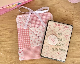 May include: A quilted fabric book sleeve featuring a pink and white patchwork pattern of gingham and floral prints, tied with a white ribbon. Beside it lies a rectangular clear plastic card holder displaying a library card design with the text "LIBRARY CARD," "THE FEATURED READERS DEPARTMENT," and "READ MORE BOOKS."