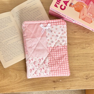 May include: A quilted fabric book sleeve featuring a patchwork design of pink and white gingham, striped, and floral patterns. The rectangular sleeve is accented with a small white ribbon bow centred near the top edge, providing a soft texture.