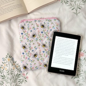 May include: A white quilted Kindle sleeve with a pink zip, patterned with bees and small blue and pink flowers. A Kindle e-reader is beside the sleeve, showing text on its screen.