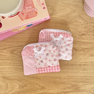 May include: Two quilted pouches featuring a patchwork design of pink gingham, pink stripes, and a white fabric with small pink and blue floral patterns. Each pouch has a white zipper closure and a small white ribbon bow attached to the front.