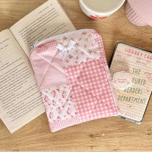 May include: A quilted fabric pouch featuring a patchwork design of pink and white gingham, striped, and floral patterns. The pouch is accented with a small white ribbon bow at the top center and includes a silver zipper closure along the top edge.