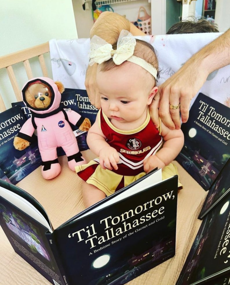 Op de afbeelding: Een babymeisje in een rood-gouden cheerleading-outfit leest een boek met de titel "'Til Tomorrow, Tallahassee: A Bedtime Story of the Garnet and Gold". Het boek is open en het meisje houdt het met beide handen vast. Er is een pluche astronautbeer op de achtergrond.