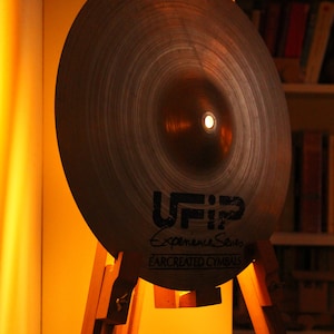Può includere: Un piatto marrone con il testo "UFIP Experience Series EARCREATED CYMBALS" su di esso, montato su un cavalletto di legno.