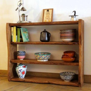 Op de afbeelding: Een houten boekenplank met drie niveaus en een natuurlijke afwerking met boomrand. De planken bevatten diverse items, waaronder boeken, een donkere keramische theepot, stapels kommen, een kom met patronen, een bloemenkan en een zwart-wit kom.