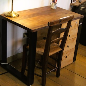 Op de afbeelding: Een houten bureau met een donkerbruin tafelblad en zwarte metalen poten. Een houten stoel met een geweven zitting staat voor het bureau. Een kleine lamp met een gouden voet staat op het bureau. Een ladekast is aan het bureau bevestigd.