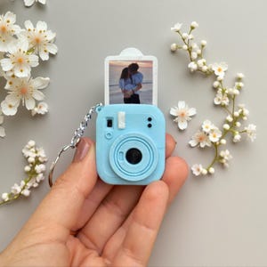 May include: A light blue camera-shaped keychain featuring a miniature instant photo print of two people embracing on a beach. The keychain includes a silver-toned metal ring and chain, designed to resemble a compact instant film camera.