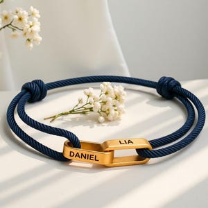 Könnte beinhalten: Ein dunkelblaues Seilarmband mit zwei ineinandergreifenden, goldfarbenen rechteckigen Metallgliedern. Die Glieder sind mit den Namen DANIEL und LIA in schwarzer Schrift graviert. Das Armband wird durch zwei verstellbare Knoten gehalten.
