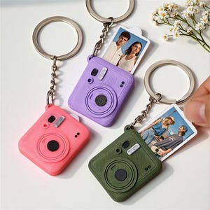 May include: Three camera-shaped keychains in pink, purple, and olive green. Each keychain features a metal ring and a small, rectangular photo insert showing people. The camera bodies have textured surfaces, a circular lens detail, and a small silver accent.