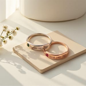 May include: Two rose gold-toned rings featuring engraved text and small inset gemstones. One ring is engraved with the name Darren and the years 1983-2015 with a green stone. The other ring is engraved with Sarah 1985-2020 with a red stone.