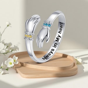 May include: A silver-toned open ring featuring two hands meeting in the centre. One hand is adorned with three yellow stones, and the other with three blue stones. The outer band is engraved with the black text Always in My heart.